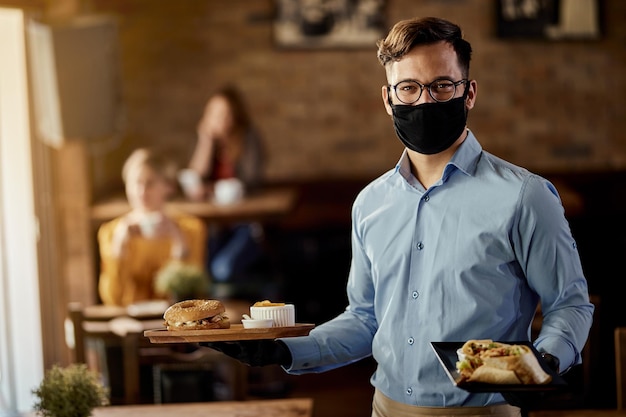 Foto gratuita joven camarero feliz con mascarilla protectora mientras sirve comida en un restaurante