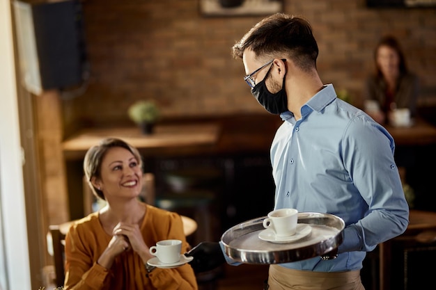 Joven camarero con mascarilla protectora mientras sirve café a un cliente en un café