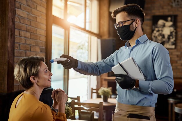 Joven camarero con mascarilla protectora y termómetro infrarrojo mientras mide la temperatura del huésped en un café