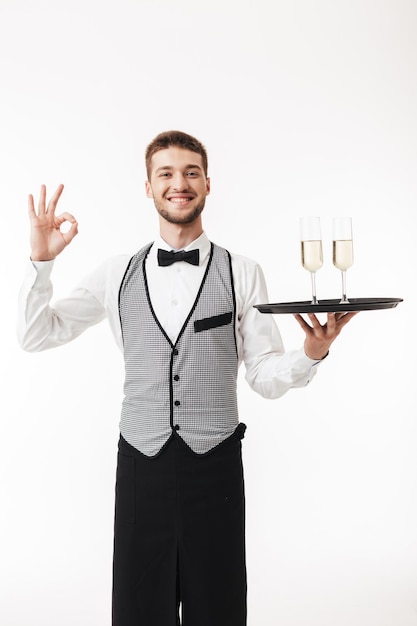 Joven camarero sonriente con uniforme mirando alegremente a la cámara sosteniendo una bandeja con gafas mientras mostraba un gesto de ok sobre fondo blanco