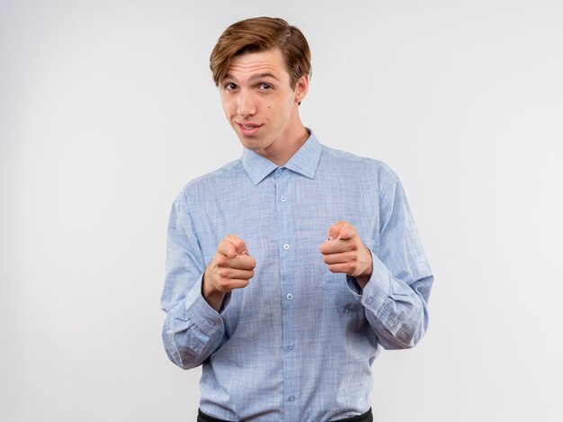 Joven con camisa azul apuntando con el dedo índice a la cámara sonriendo confiada feliz y positiva de pie sobre fondo blanco.