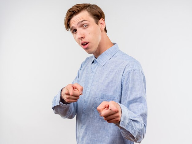 Joven con camisa azul apuntando con los dedos índices sonriendo confiado de pie sobre la pared blanca