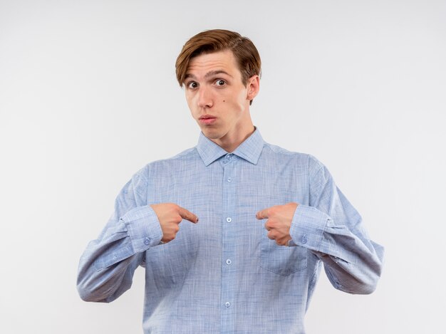 Joven con camisa azul apuntando a sí mismo confundido y sorprendido de pie sobre la pared blanca