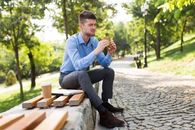 Joven con camisa azul comiendo sándwich con una taza de café para llevar y una computadora portátil cerca de un banco en el parque de la ciudad verde