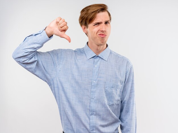 Joven con camisa azul disgustado mostrando los pulgares hacia abajo de pie sobre la pared blanca