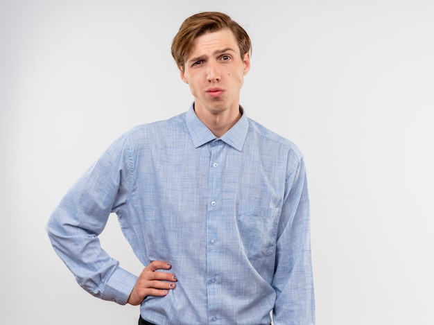 Foto gratuita joven con camisa azul mirando a la cámara con la mano en la cadera mirando con expresión escéptica de pie sobre fondo blanco.