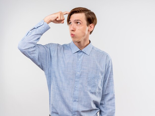 Joven con camisa azul mirando a un lado desconcertado rascándose la cabeza de pie sobre la pared blanca