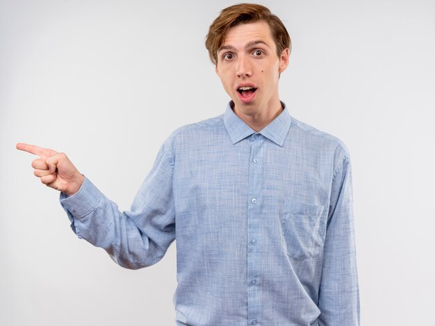 Joven en camisa azul pointign con el dedo índice hacia el lado sonriendo alegremente de pie sobre fondo blanco.