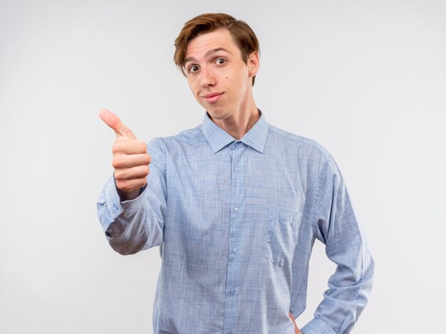 Joven en camisa azul sonriendo mostrando los pulgares para arriba de pie sobre la pared blanca