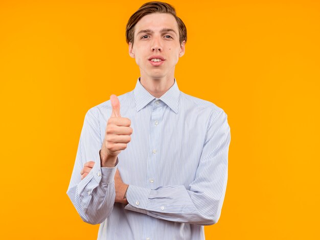 Joven en camisa blanca sonriendo confiado mostrando Thumbs up parado sobre pared naranja