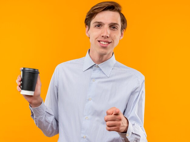 Joven en camisa blanca sosteniendo la taza de café apuntando con el dedo índice sonriendo confiado de pie sobre la pared naranja