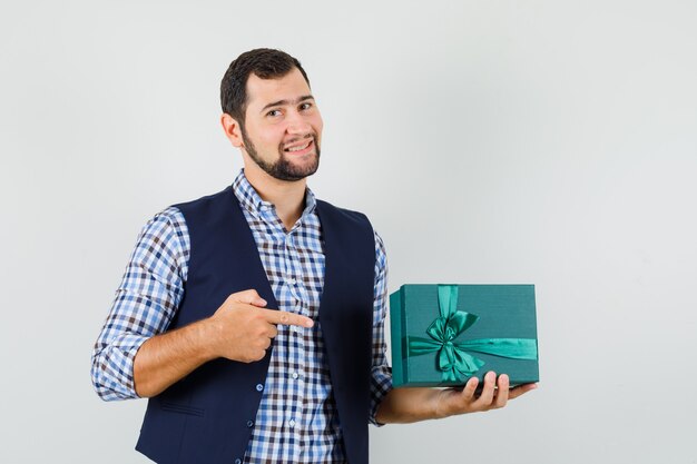 Joven en camisa, chaleco apuntando a la caja actual y mirando jovial, vista frontal.