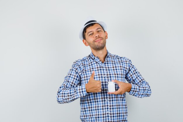 Joven en camisa de cuadros, sombrero sosteniendo una botella de píldoras