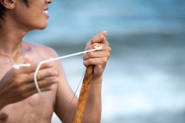 Joven sin camisa en la playa con arco de madera preparándose para practicar capoeira