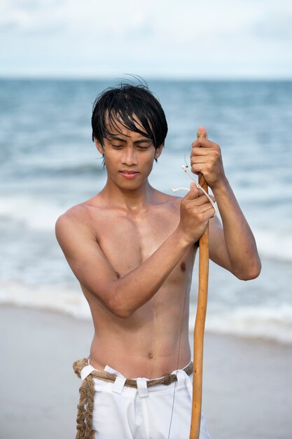 Joven sin camisa en la playa con arco de madera preparándose para practicar capoeira