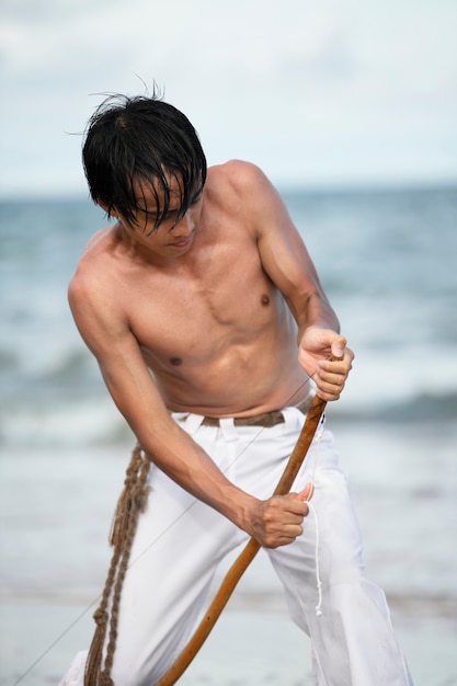 Foto gratuita joven sin camisa en la playa con arco de madera preparándose para practicar capoeira