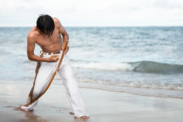 Joven sin camisa en la playa con arco de madera preparándose para practicar capoeira