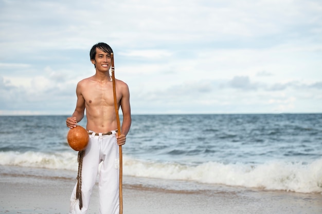 Foto gratuita joven sin camisa en la playa con arco de madera preparándose para practicar capoeira