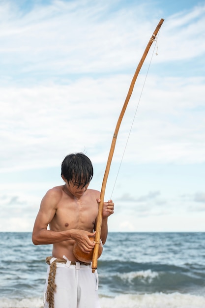 Foto gratuita joven sin camisa en la playa con arco de madera preparándose para practicar capoeira