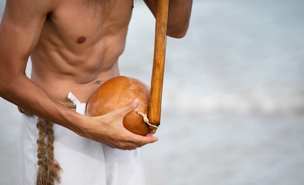 Joven sin camisa en la playa con arco de madera preparándose para practicar capoeira