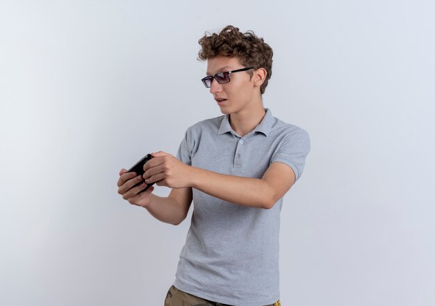 Joven en camisa de polo gris mirando la pantalla de su teléfono inteligente jugando juegos de pie sobre la pared blanca