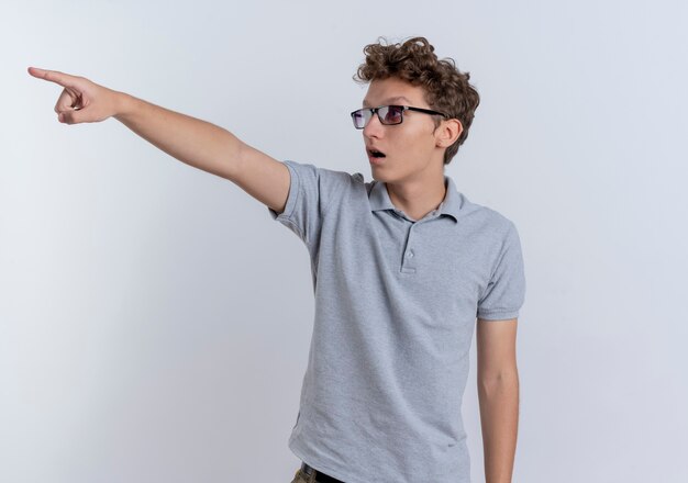 Joven en camisa polo gris mirando sorprendido apuntando con el dedo índice a algo parado sobre la pared blanca