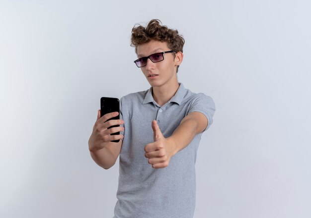 Joven en camisa de polo gris con smartphone mostrando pulgares para arriba de pie sobre la pared blanca