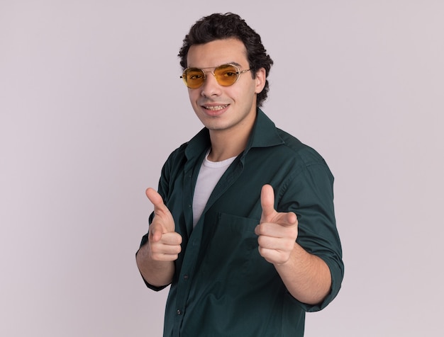 Joven en camisa verde con gafas apuntando con los dedos índices al frente feliz y positivo de pie sobre la pared blanca