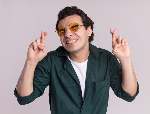 Joven en camisa verde con gafas loco feliz cruzando los dedos con los ojos cerrados de pie sobre la pared blanca