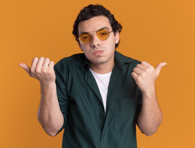 Joven en camisa verde con gafas mirando al frente confundido soplando mejillas apuntando con los pulgares a los lados de pie sobre la pared naranja