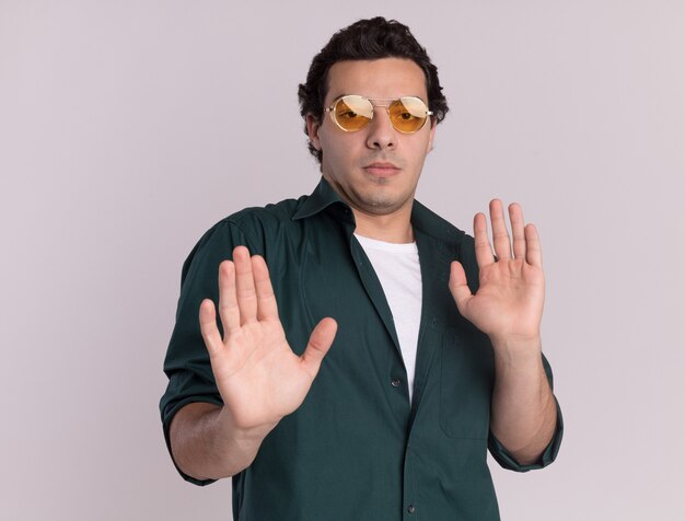 Joven en camisa verde con gafas mirando al frente preocupado haciendo gestos de defensa con las manos de pie sobre la pared blanca