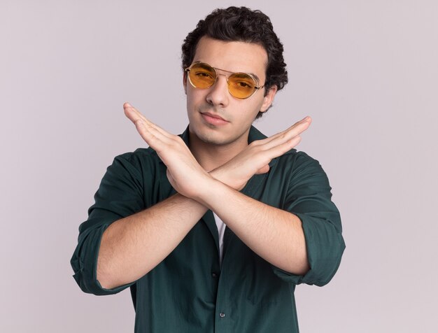 Joven en camisa verde con gafas mirando al frente con rostro serio cruzando las manos de pie sobre la pared blanca