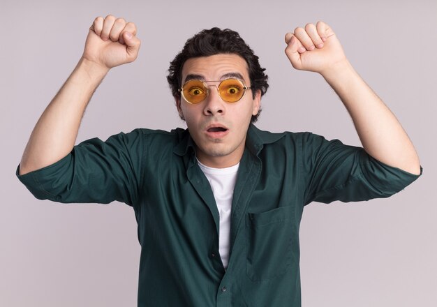 Joven en camisa verde con gafas mirando al frente sorprendido y asombrado levantando los puños de pie sobre la pared blanca