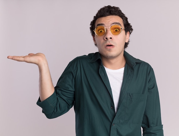 Joven en camisa verde con gafas mirando al frente sorprendido presentando copia espacio con el brazo de la mano de pie sobre la pared blanca