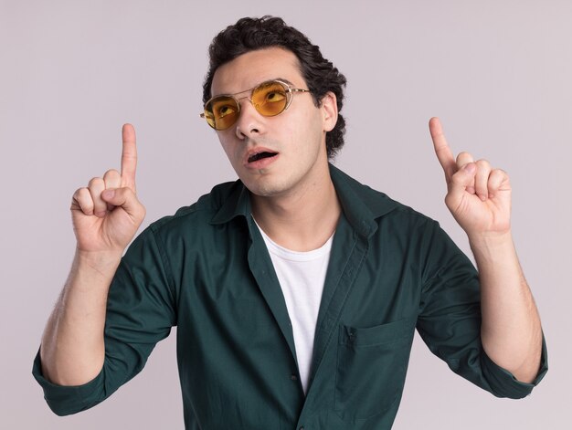 Joven en camisa verde con gafas mirando hacia arriba desconcertado apuntando con los dedos índices de pie sobre la pared blanca