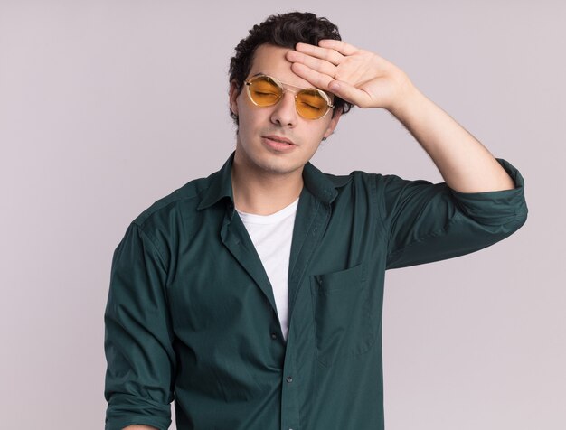 Joven en camisa verde con gafas mirando cansado y aburrido con la mano en la frente de pie sobre la pared blanca