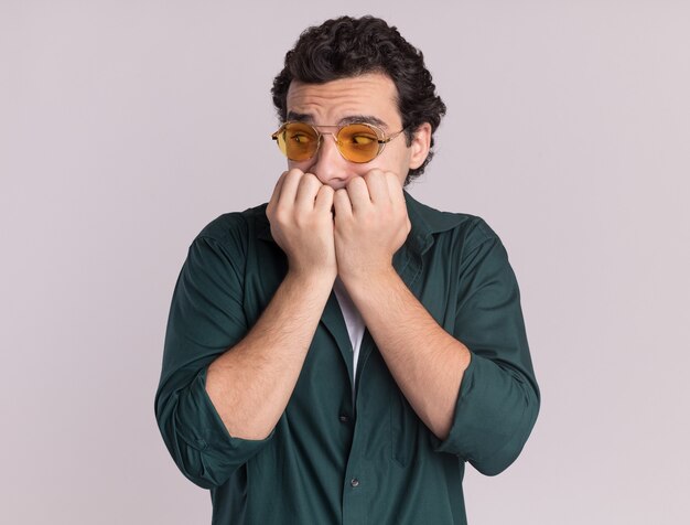 Joven en camisa verde con gafas mirando a un lado estresado y nervioso morderse las uñas de pie sobre la pared blanca