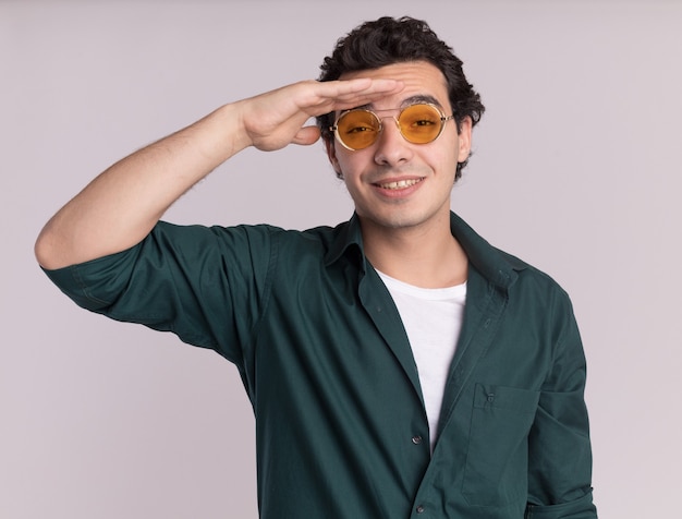 Joven en camisa verde con gafas mirando a lo lejos con las manos sobre la cabeza de pie sobre la pared blanca