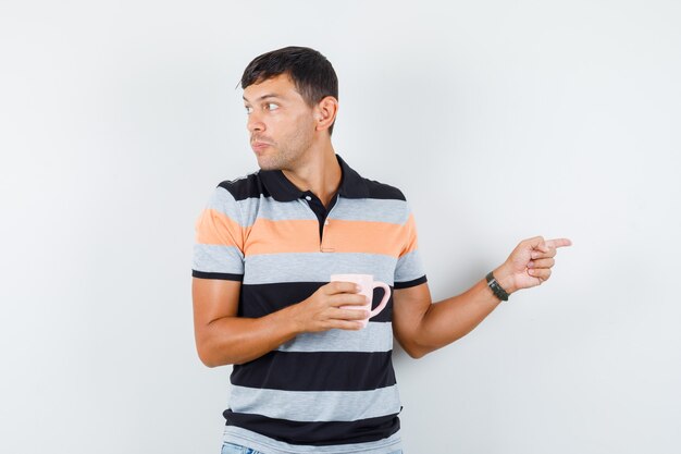 Joven en camiseta apuntando hacia afuera mientras sostiene una taza de bebida y mirando enfocado