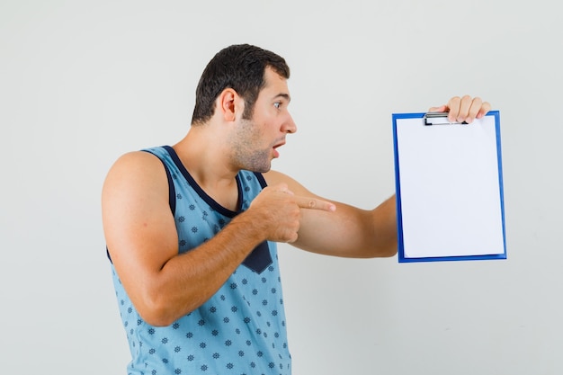 Foto gratuita joven en camiseta azul apuntando al portapapeles y mirando sorprendido