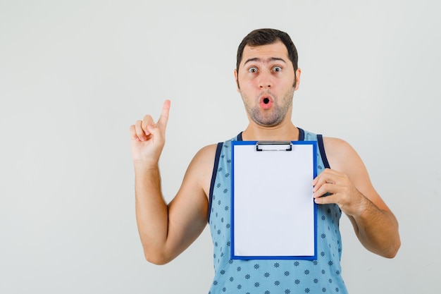 Joven en camiseta azul apuntando hacia arriba mientras sostiene el portapapeles y parece sorprendido