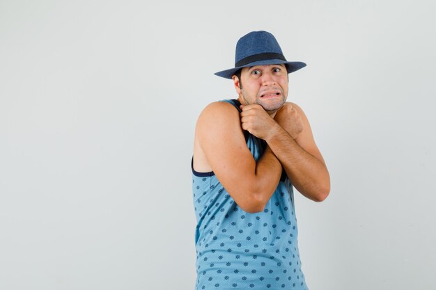 Joven en camiseta azul, sombrero sosteniendo puños cruzados en el pecho y mirando indefenso