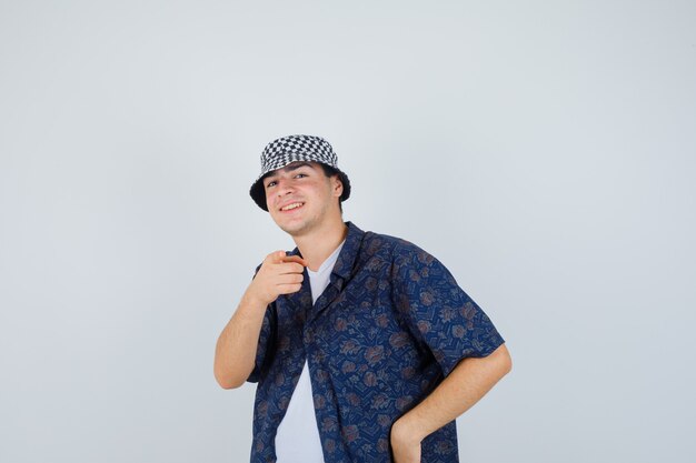 Joven con camiseta blanca, camisa floral, gorra apuntando a la cámara con el dedo índice y mirando feliz, vista frontal.