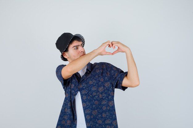 Joven en camiseta blanca, camisa floral, gorra mostrando gesto de corazón y mirando confiado