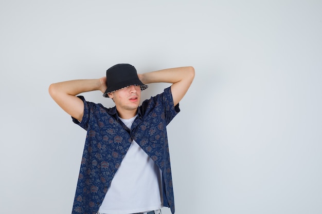 Joven con camiseta blanca, camisa floral, gorra sosteniendo las manos detrás de la cabeza y mirando confiado, vista frontal.