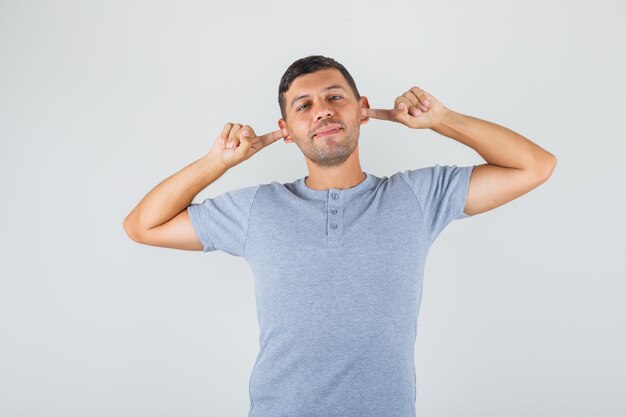 Joven en camiseta gris cubriendo las orejas con los dedos y mirando contento