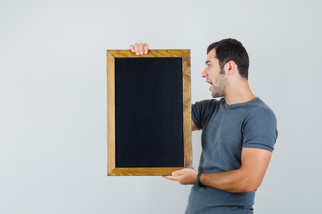 Foto gratuita joven en camiseta gris sosteniendo pizarra y mirando enfocado