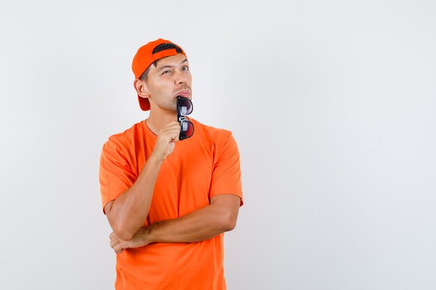 Joven en camiseta naranja y gorra sosteniendo vasos y mirando vacilante