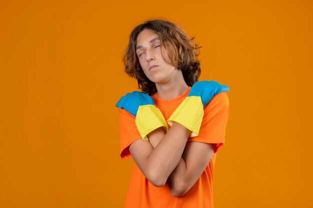 Joven en camiseta naranja con guantes de goma abrazándose a sí mismo con los ojos cerrados de pie sobre fondo amarillo