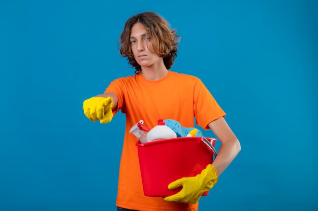 Joven en camiseta naranja con guantes de goma sosteniendo un balde con herramientas de limpieza apuntando a la cámara de pie disgustado sobre fondo azul.